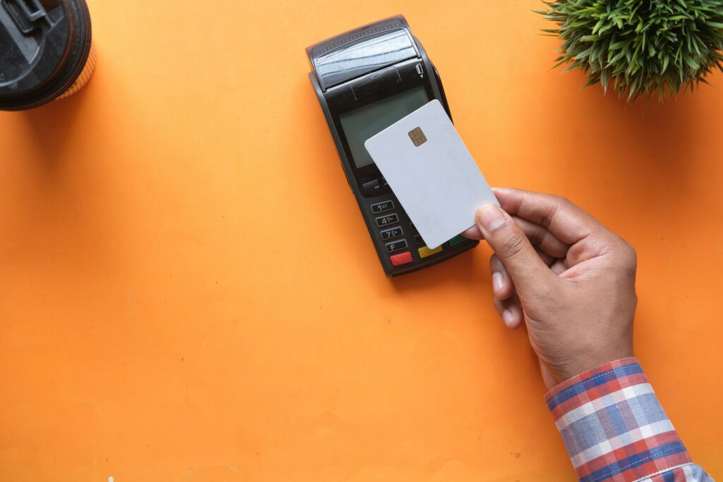 Persons hand paying with a credit card over a light orange background with a green plant in the top right corner